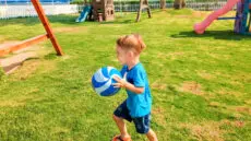 Portrait of happy smiling little boy holding ball in hands and running on grass at children playground